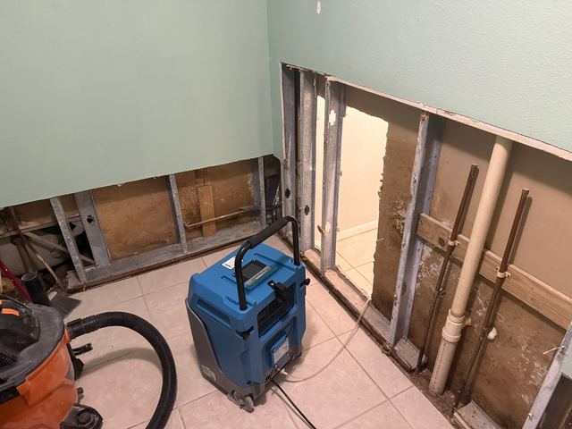 Mold-stained wall cavity with pipes in an opened bathroom wall, and the same bathroom with drying equipment running.